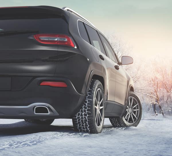 A sleek black SUV parked on a snowy road, showcasing winter tires with a tread pattern designed for traction on icy surfaces. The background features snow-covered trees under a soft, warm sunrise or sunset glow.