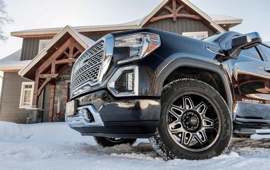 A  close-up of a black pickup truck with Hercules Tires’ Avalanche TT winter tires parked in front of a snow-covered house, showcasing the rugged tread pattern designed for extreme winter conditions.