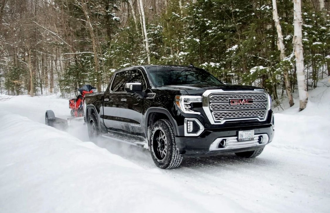 A black pickup truck with Hercules Tires’ Avalanche TT winter tires towing a red snowmobile on a snow-covered road in a woodsy area.